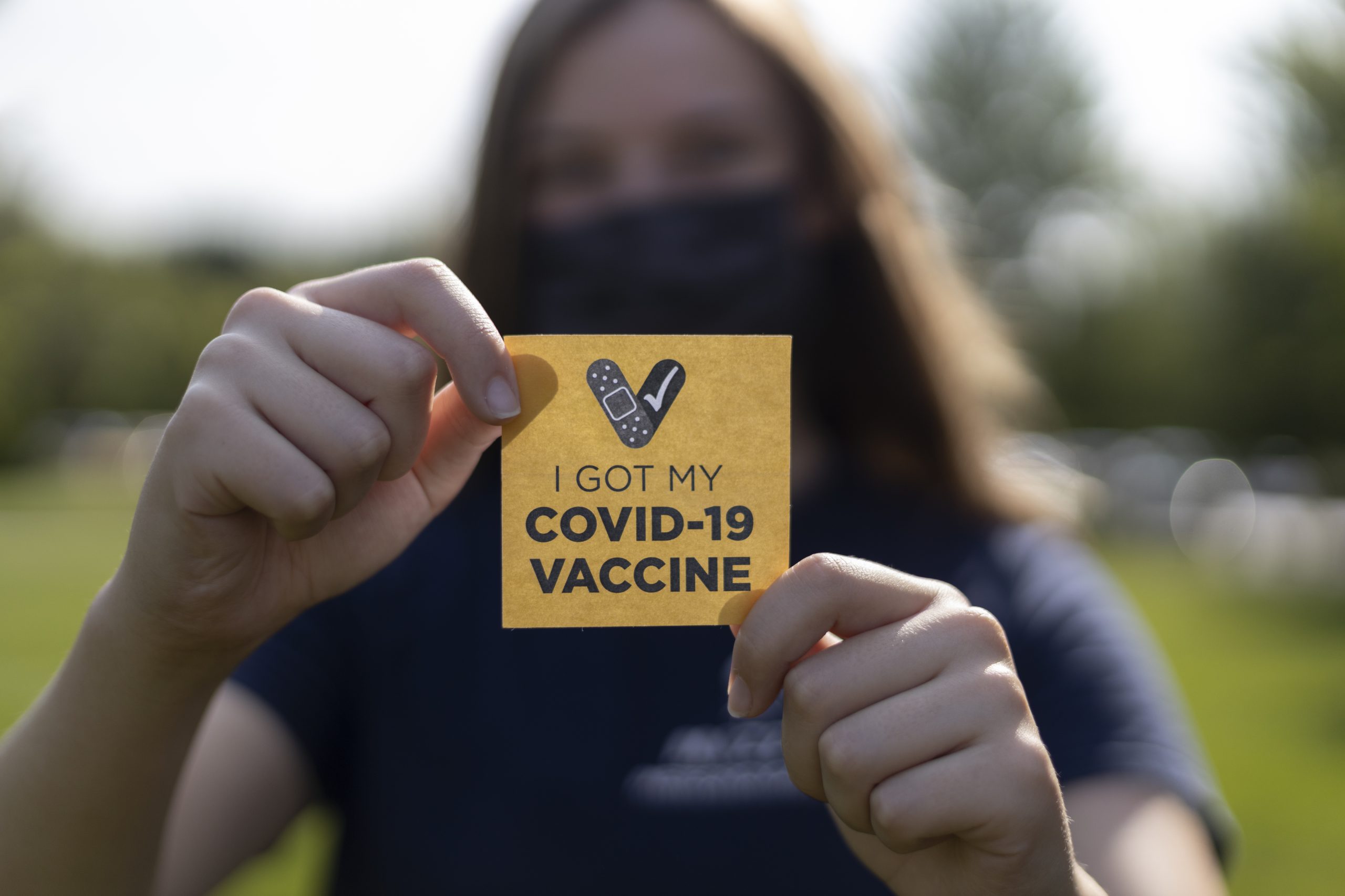 Masked student holds a sticker promoting COVID-19 vaccine
